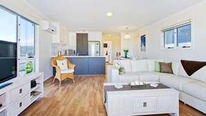 A modern living room with a white sofa, a coffee table, and a TV on a wooden cabinet. There is a kitchen area with a fridge, microwave, and a dining table with chairs. - Sunset at Snapper Point ~ Aldinga Beach - Modern Coastal Beauty with Large Balcony and Sea Views (Aldinga Beach)