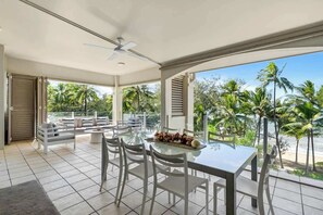Uma ampla área de refeições ao ar livre com mesa e cadeiras de vidro, ventilador de teto e vista para palmeiras e praia.