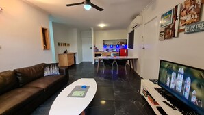 Una sala de estar moderna con un sofá de cuero marrón, una mesa de centro blanca y un televisor de pantalla plana. Se aprecia una zona de cocina con una mesa de comedor y sillas al fondo.