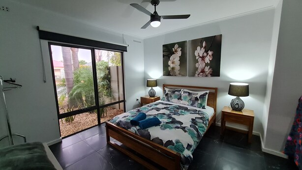 Un dormitorio con cama, dos mesitas de noche, ventilador de techo y ventana con vista a árboles y edificios.