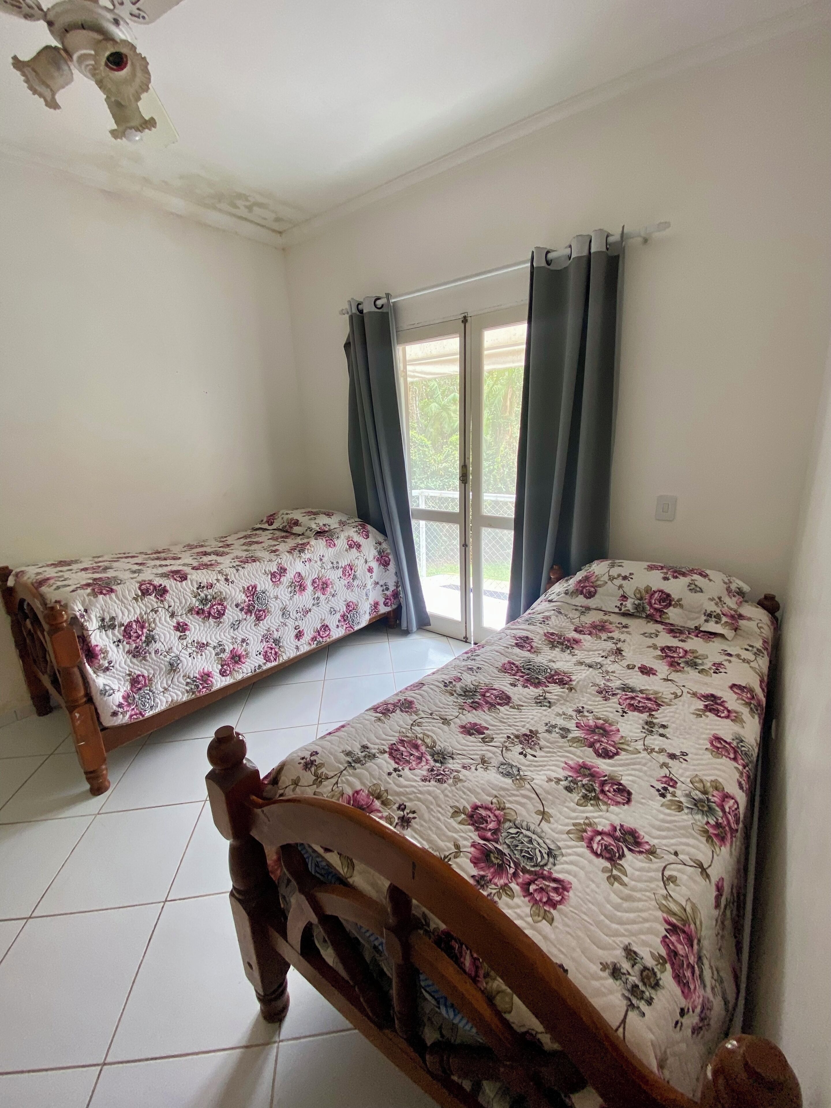 A room with two wooden beds, each with floral bedding, a tiled floor, and a window with curtains.