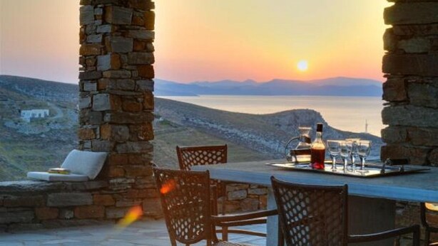 Ein Steintisch mit Stühlen und einer Weinflasche, mit Blick auf eine malerische Wasserlandschaft und Berge bei Sonnenuntergang.