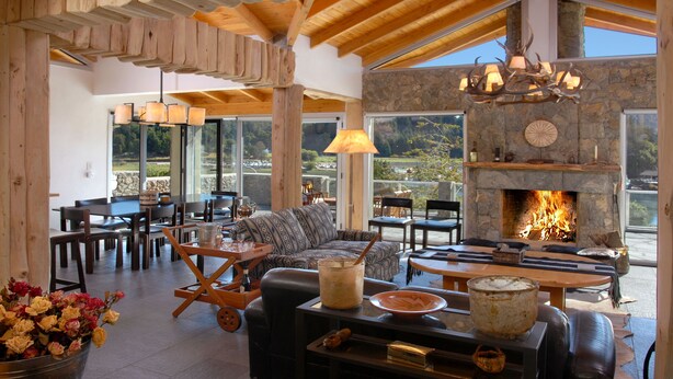 Un amplio salón con chimenea de piedra, muebles de madera y grandes ventanales con vistas al exterior.