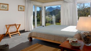 Un dormitorio con una cama, una silla de madera, una mesa con lámpara y vistas a montañas a través de la ventana.