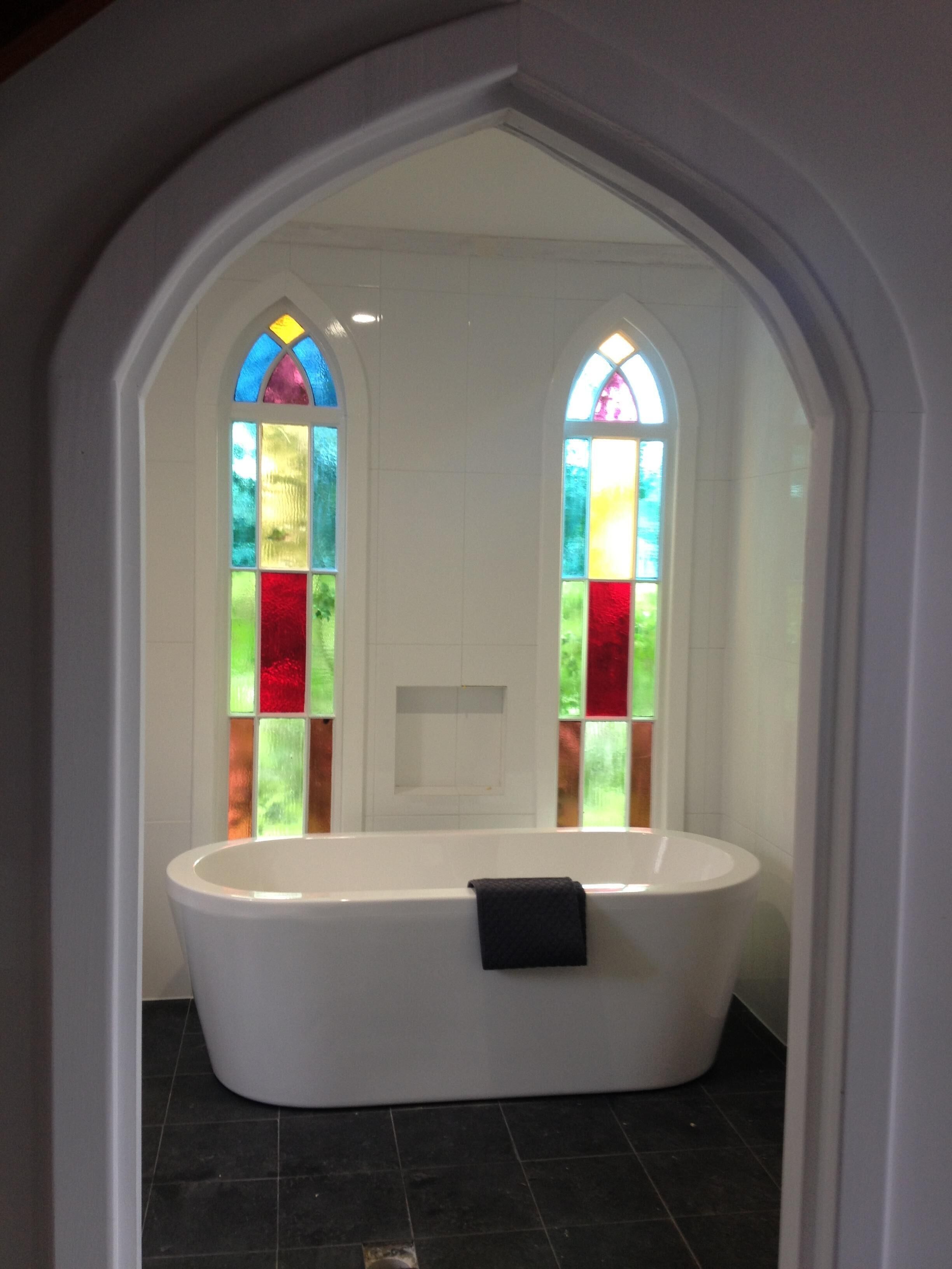 A bathroom with a freestanding bathtub, two stained glass windows, and a white tiled wall.