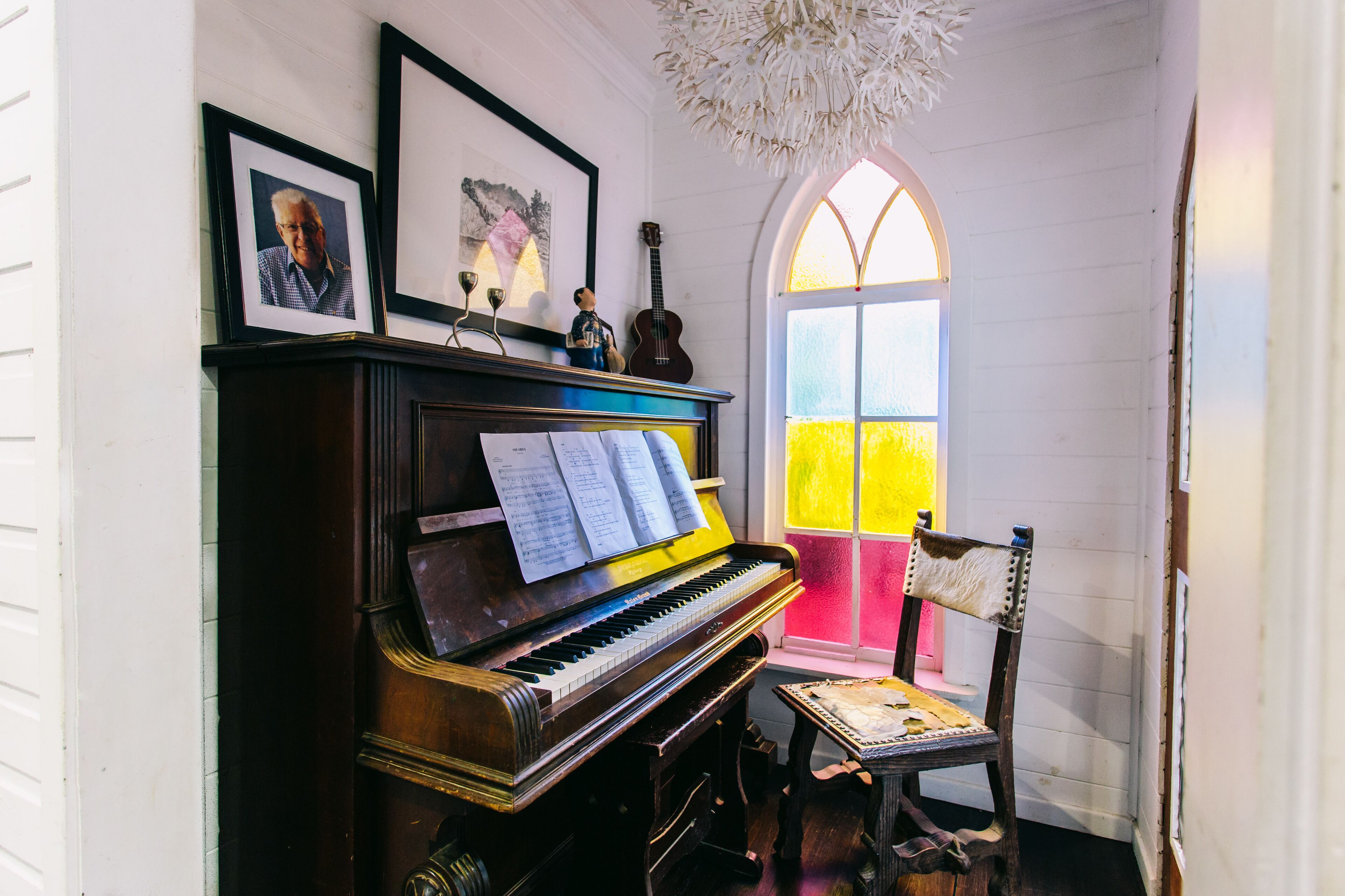 A piano with sheet music, a framed photograph, a guitar, and a chair in a room with white walls and a stained glass window.