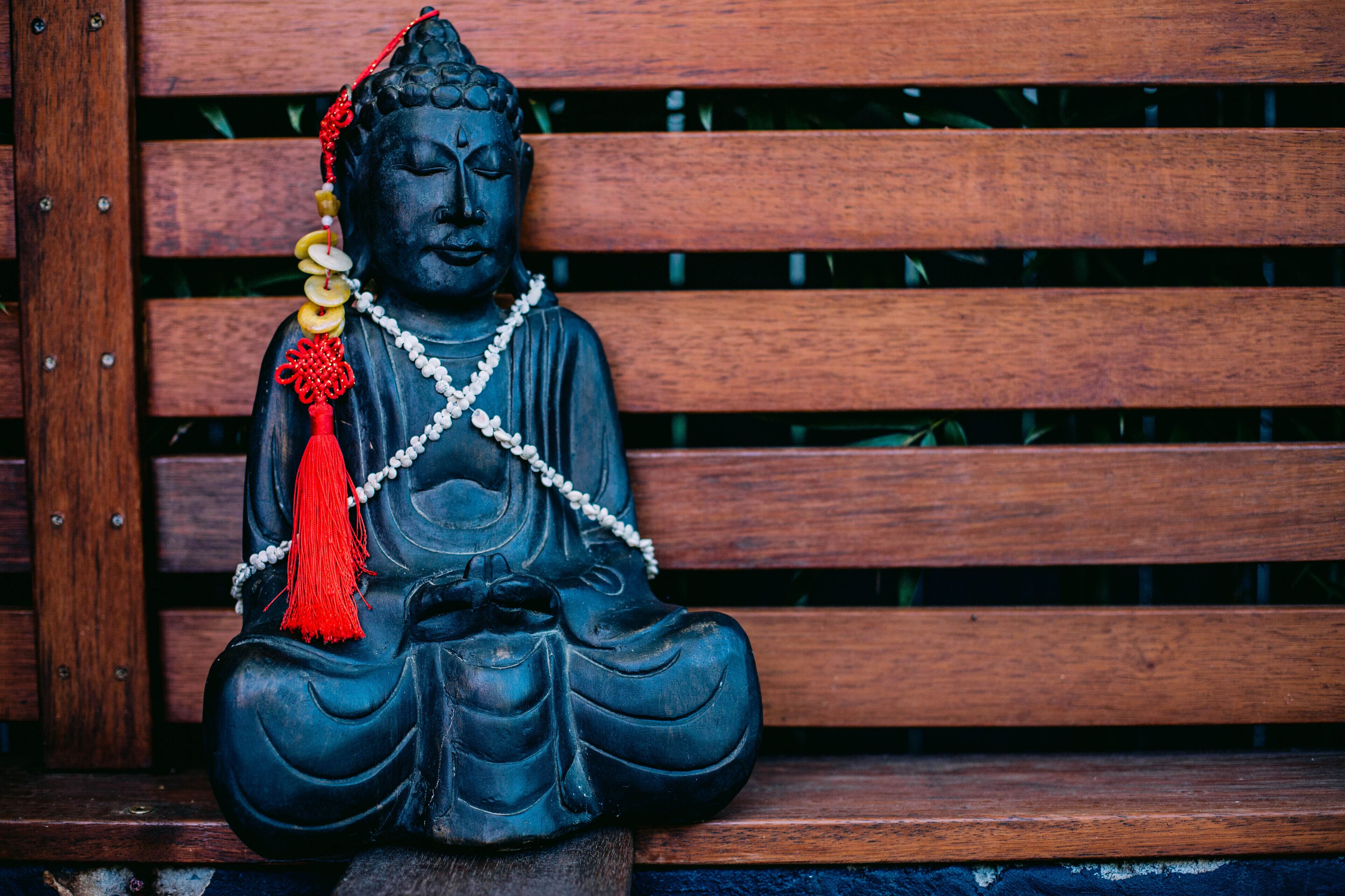 A statue of Buddha with a red and white beaded necklace, seated on a wooden bench.