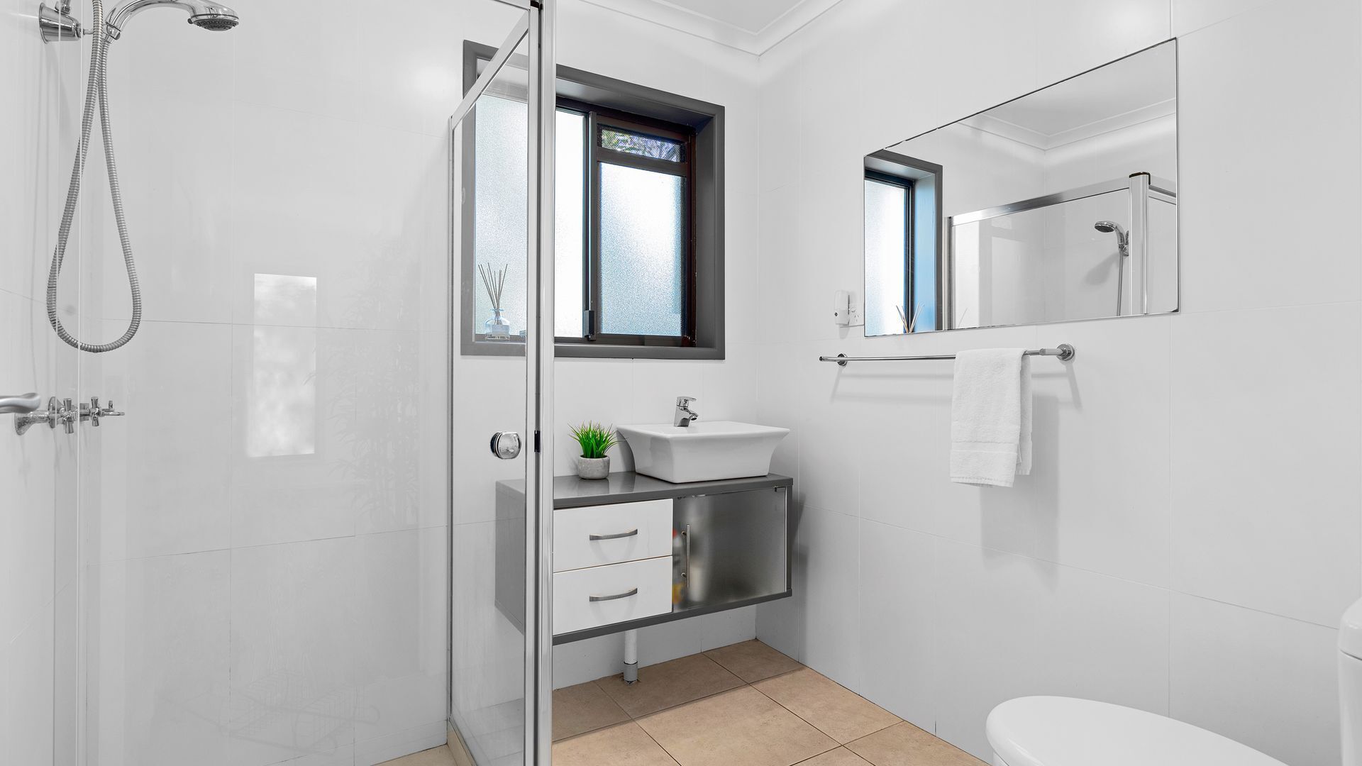 A modern bathroom with a glass shower enclosure, a sink with a small plant, a window, and a toilet.