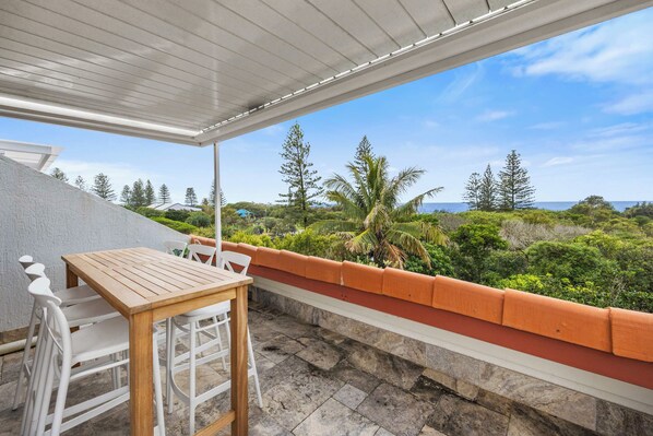A covered outdoor seating area with a table and chairs, overlooking a lush landscape with trees and a view of the sea. - Peregian Beach Apartment (Peregian Beach)
