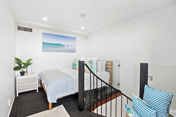 Een slaapkamer met een bed, een nachtkastje, een kamerplant, een schilderij van een strand en een trap met een leuning.