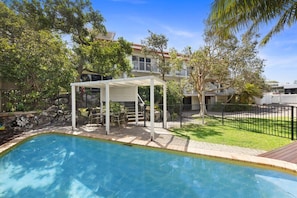 A swimming pool with a covered seating area, a fenced yard, and a building with a balcony in the background.
