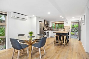 A modern kitchen with a central island, a dining area with a glass table, and a view of a garden through large windows. - Peregian Beach Apartment (Peregian Beach)