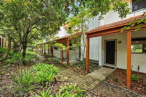 A covered outdoor area with a pathway leading to a building entrance, surrounded by greenery and plants.