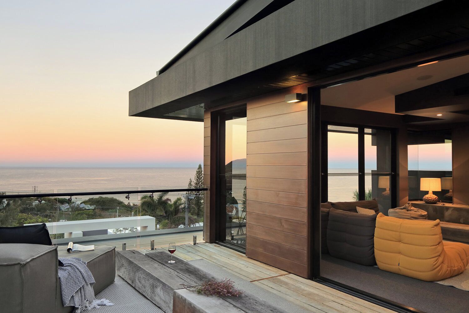 A modern balcony with a view of the ocean at sunset, featuring a sofa and a glass door leading to an interior space.