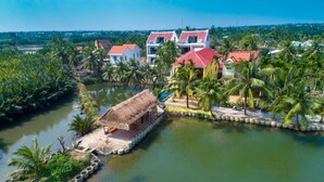 A riverside property with a thatched-roof structure, a pool, and several multi-story buildings with red roofs.
