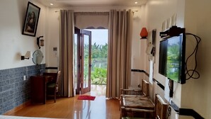 A room with a wooden floor, a TV mounted on the wall, a dining table with chairs, and a view of greenery outside.