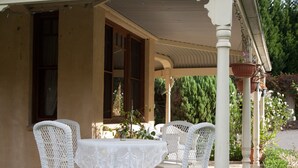 A covered outdoor seating area with white wicker furniture and a table, surrounded by greenery and a potted plant.