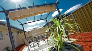 En udendørs terrasse med pergola, siddepladser og en potteplante.