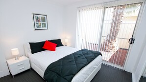 Et hotelværelse med seng, et natbord, en lampe, et billede i ramme på væggen og udsigt udad gennem en dør med metalgitter.
