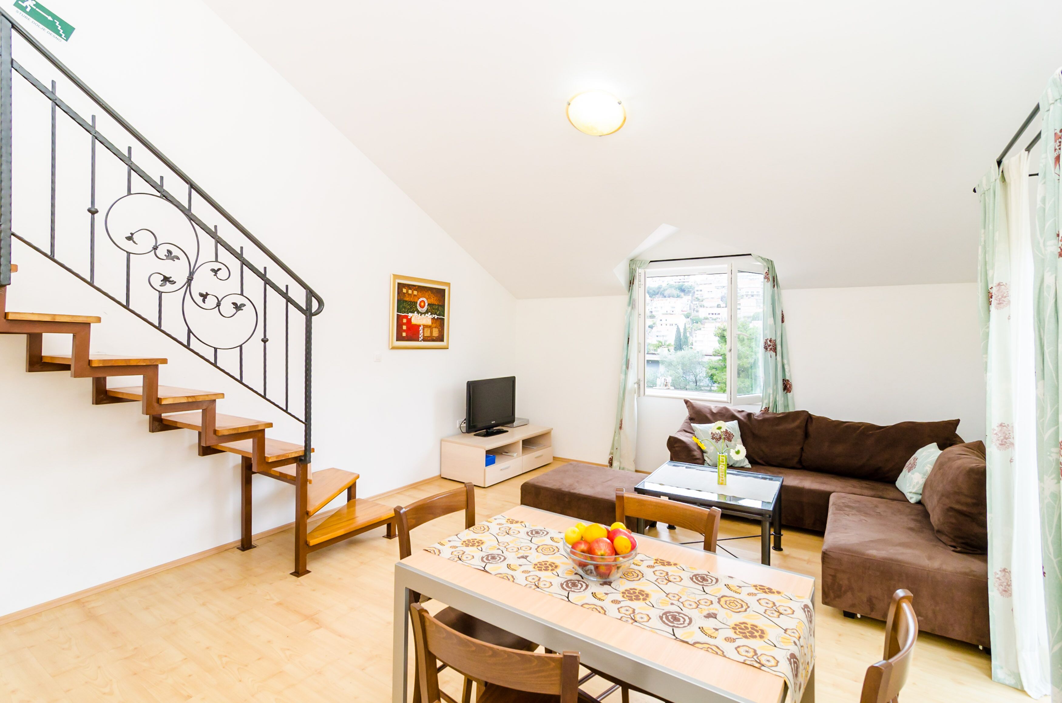 Un salon avec un escalier, une table à manger avec des chaises, un canapé, un téléviseur posé sur un meuble et une fenêtre avec des rideaux.