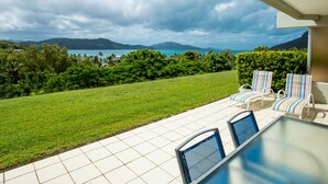 A patio with two striped lounge chairs and a table, overlooking a lush green lawn and a scenic view of a body of water and mountains.