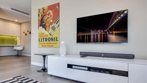 Een moderne woonkamer met een flatscreen televisie aan de muur, een witte entertainmentsunit eronder, en een poster van een vrouw die een fles Citronil vasthoudt.