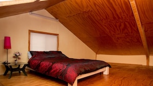 Ein Schlafzimmer mit schrägen Wänden, einem Bett mit gemustertem Überwurf, einem Nachttisch mit Lampe und einem Fenster mit Jalousien.