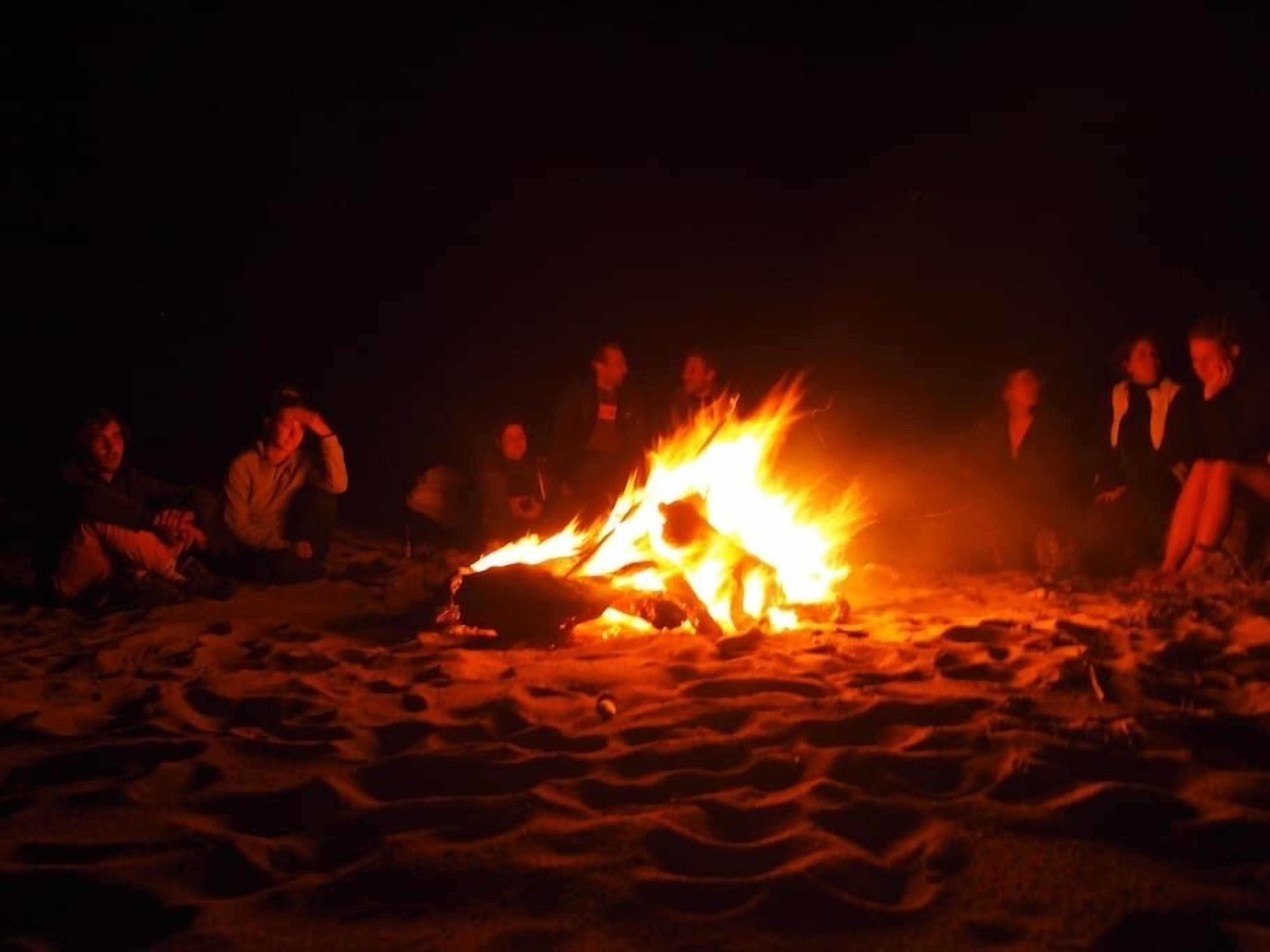 A group of people sitting around a campfire on a sandy beach at night.