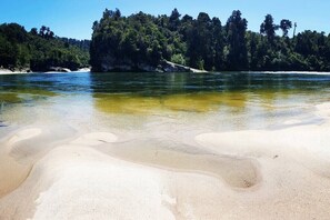 Orilla de río con agua cristalina, playa de arena y un área boscosa al fondo.