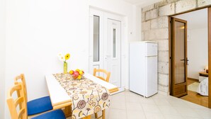 Ein kleiner, heller Raum mit einer weißen Tür, einem weißen Kühlschrank, einem Tisch mit gemustertem Tischläufer und einem Stuhl mit blauer Sitzkissen.