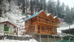 A wooden multi-story building with balconies, surrounded by snow-covered trees and a stone wall. - Lato The Huts River Touch Guesthouse  (Kullu)