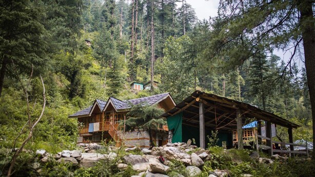 A rustic cabin with a wooden exterior, a stone foundation, and a green roof, surrounded by dense forest and rocky terrain. - Lato The Huts River Touch Guesthouse  (Kullu)