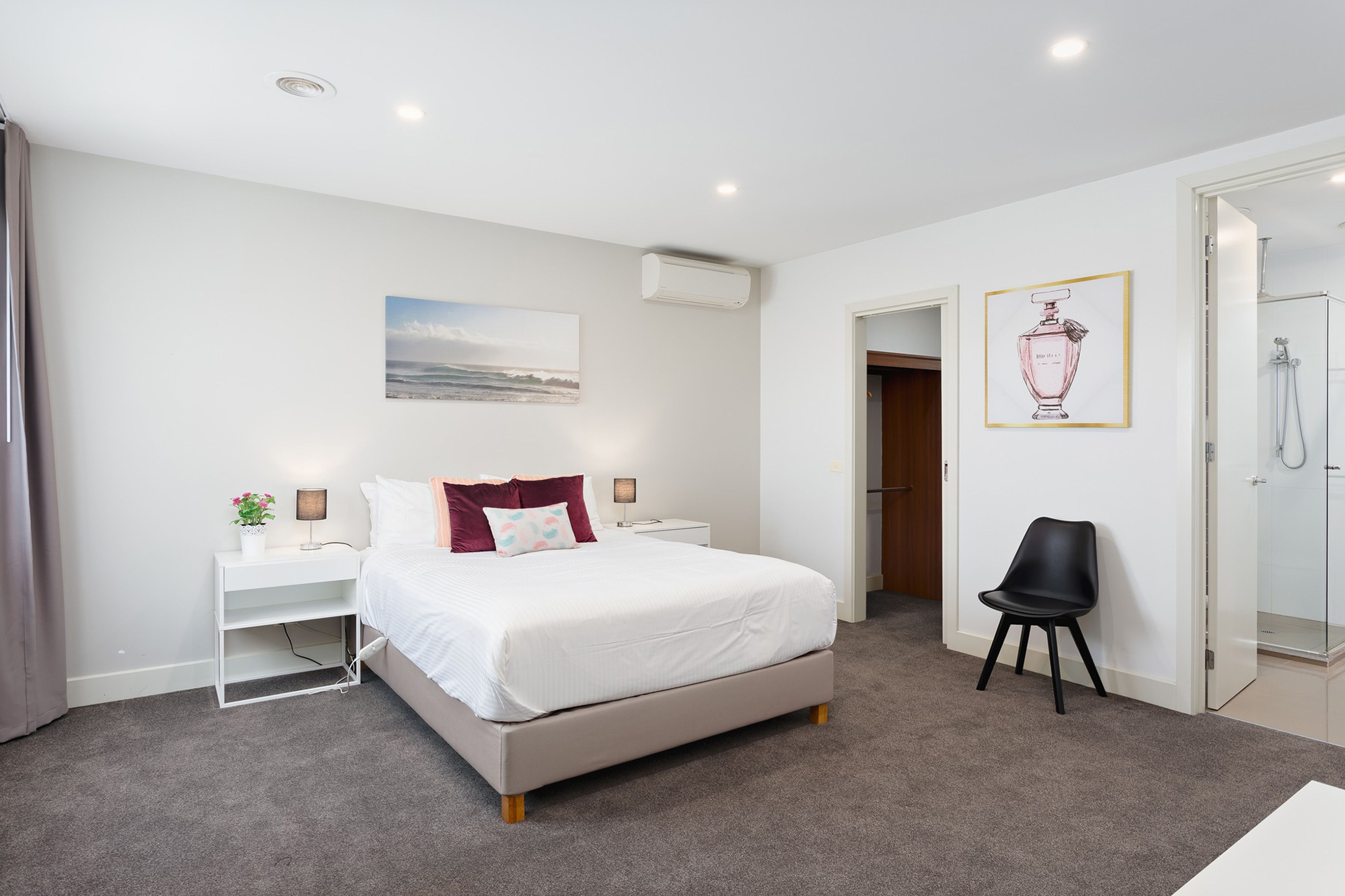 A modern hotel room with a bed, bedside tables, a desk, a chair, a shower area, and a framed picture on the wall.