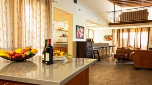 A kitchen with a fruit bowl, wine bottles, and a dining area with a sofa and a balcony. - Villa Nasia (Neo Chorio)