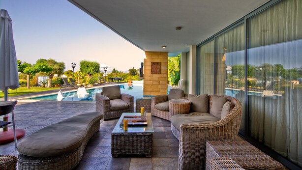A covered outdoor seating area with wicker furniture, a pool, and a view of the surrounding landscape. - Villa Nasia (Neo Chorio)