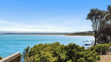 Vistes costaneres amb una terrassa de fusta que domina el mar, una platja sorrenca i vegetació abundant.