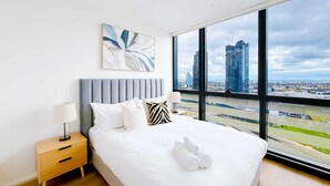A modern hotel room with a large bed, a bedside table with a lamp, a city view through large windows, and a piece of abstract art on the wall.
