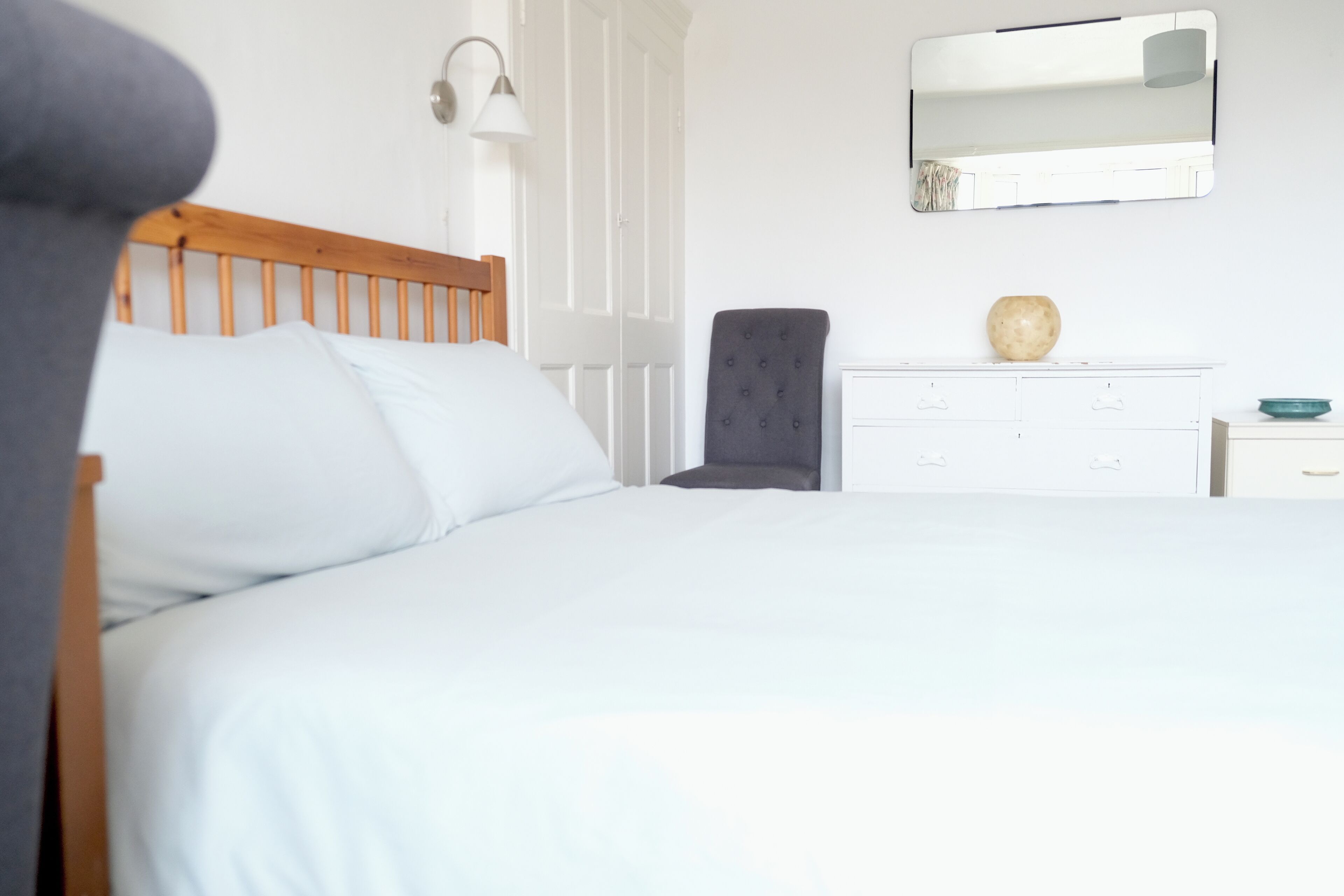 Uma cama bem arrumada com lençóis brancos, cabeceira de madeira, luminária de cabeceira, poltrona escura com tecido acolchoado, cômoda branca com um vaso e um espelho na parede.