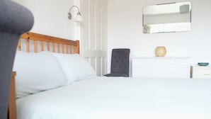Ein ordentlich bezogenes Bett mit weißen Leinen, ein hölzernes Kopfteil, eine Nachttischlampe, ein dunkel gepolsterter Sessel, eine weiße Kommode mit einer Vase und ein Spiegel an der Wand.