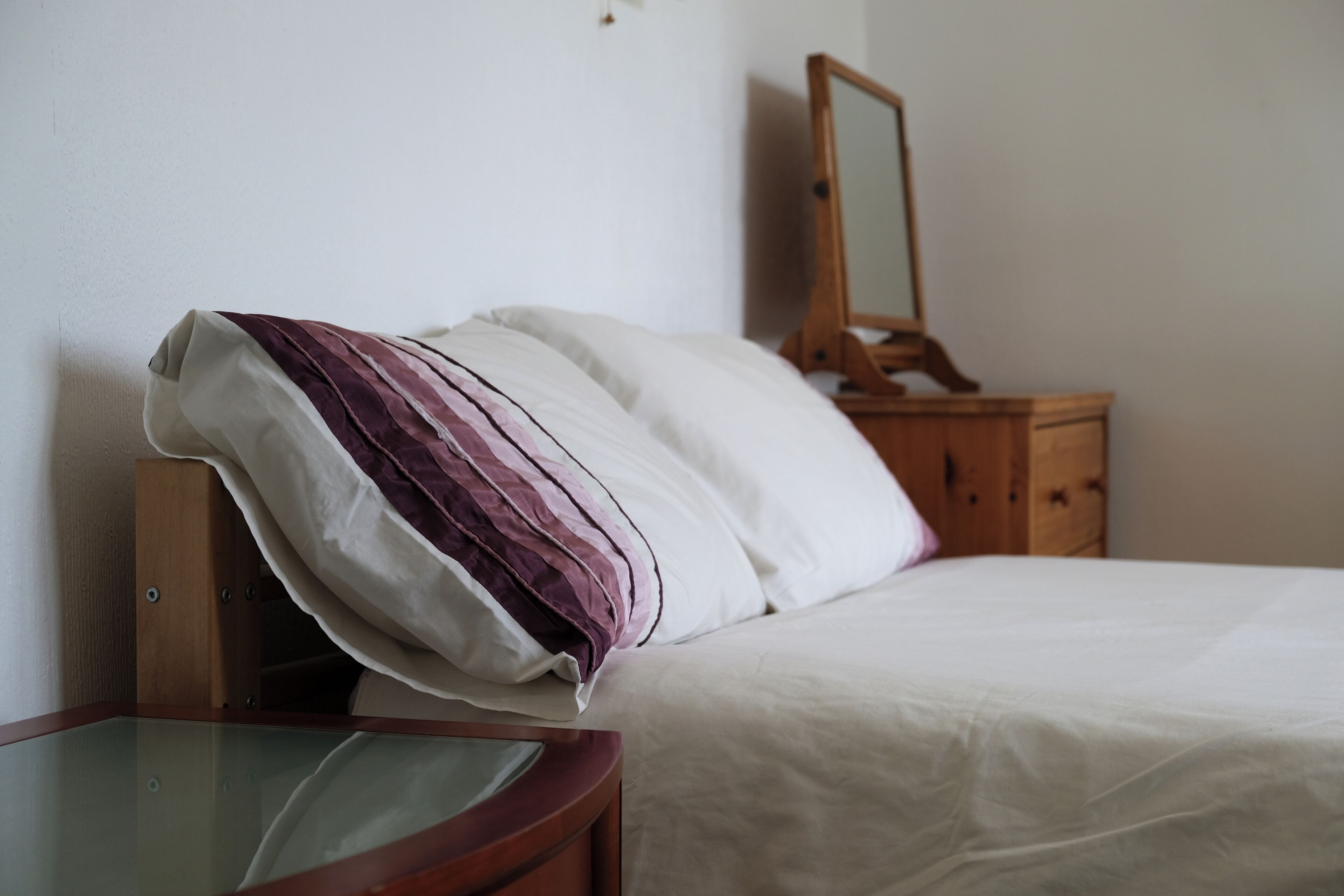 Uma cama bem arrumada com almofadas listradas em branco e roxo, uma mesa de cabeceira de madeira com um espelho e uma mesa de vidro em um quarto simples.