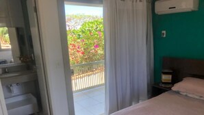 A bedroom with a bed, a nightstand, a window with curtains, and a view of a balcony with plants. - House in Buzios Geribá  (Armação dos Búzios)
