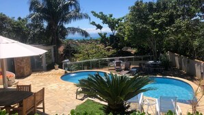 A swimming pool with lounge chairs and a table, surrounded by greenery and a view of the ocean. - House in Buzios Geribá  (Armação dos Búzios)