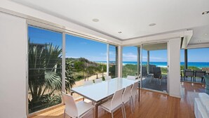 A spacious room with a large table and chairs, large windows offering a view of the beach and ocean, and a balcony with outdoor seating. - The Pavillion 1 - Beachfront (Wamberal)
