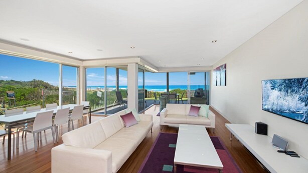 A modern living room with a large sofa, a coffee table, and a TV on a stand. There are large windows offering a view of the ocean and greenery. - The Pavillion 1 - Beachfront (Wamberal)