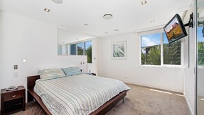 A modern bedroom with a large bed, a bedside table, a wall-mounted TV, and a window with a view of greenery. - The Pavillion 1 - Beachfront (Wamberal)