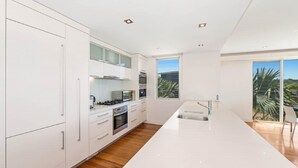 A modern kitchen with white cabinetry, a central island, and a large window providing a view of palm trees. - The Pavillion 1 - Beachfront (Wamberal)