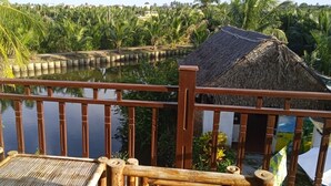 Une terrasse en bois dotée d’une balustrade, qui offre une vue sur une étendue d’eau, et qui est entourée d’une végétation luxuriante et d’une structure au toit de chaume.