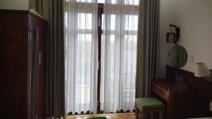 Ein Hotelzimmer mit einem Holzschrank, einem Schreibtisch mit Lampe, einem grün gepolsterten Sessel und einem großen Fenster mit durchsichtigen Vorhängen.