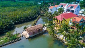 Une maison dotée d’une toiture de chaume, située à proximité d’un plan d’eau, et entourée d’une végétation luxuriante et de palmiers.