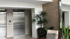 Um lobby moderno com design em dois andares, apresentando um elevador central, área de estar com uma cadeira e uma mesinha, e plantas em vasos.